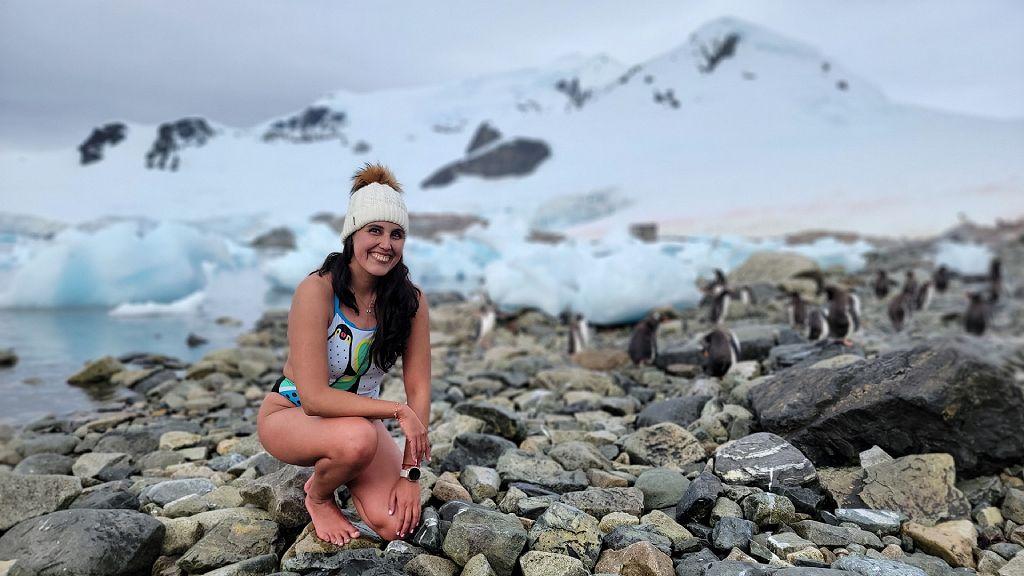 Bárbara en vestido de baño y un gorro, en una playa de Antártica con rocas y pingüinos.