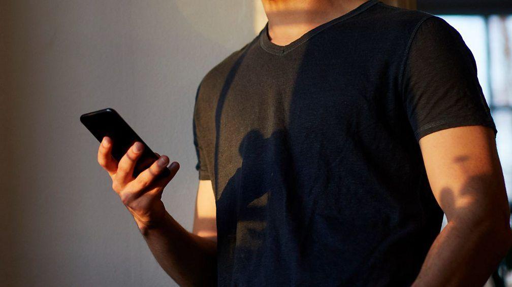 Un hombre joven con camiseta oscura de manga corta, enviando textos en su teléfono inteligente