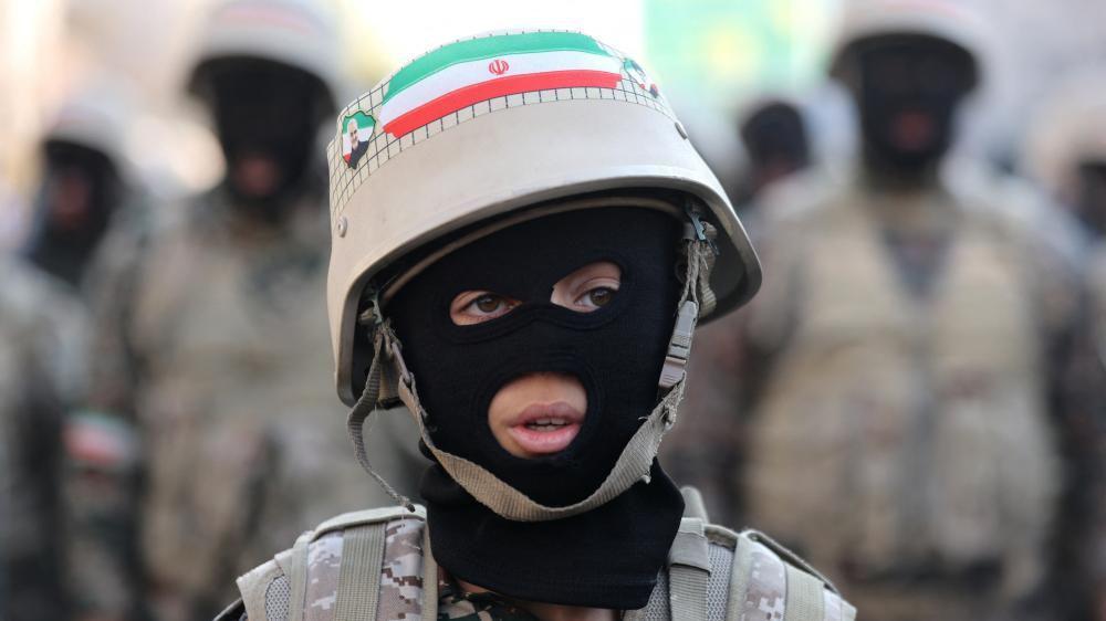Un joven que lleva un pasamontañas, uniforme de fatiga y un casco con una bandera iraní mira fuera de cámara, con figuras militares borrosas en el fondo.