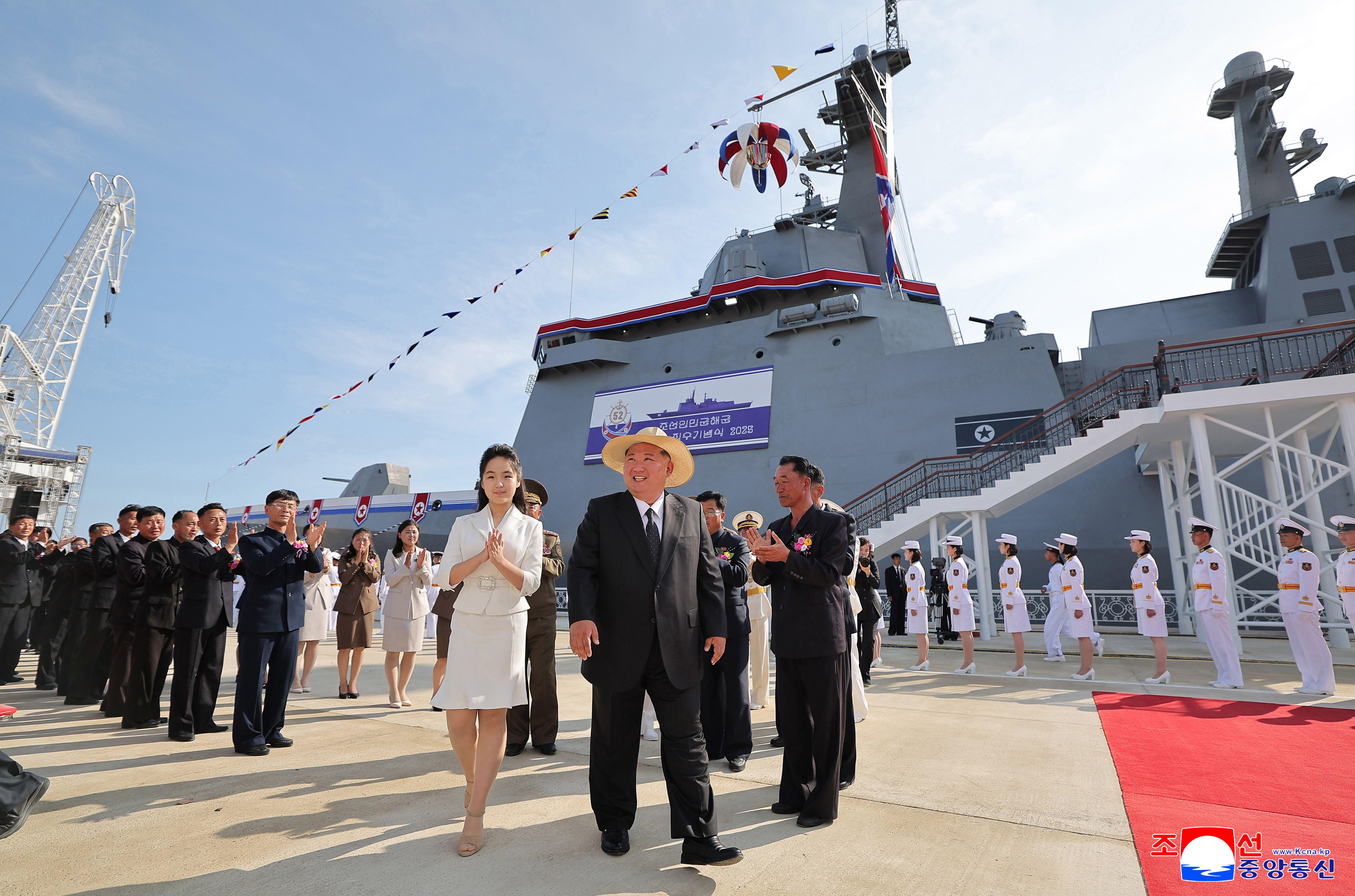 Kim Jong Un, con traje y sombrero de paja, sonríe y mira de reojo mientras camina junto a su hija, que viste americana y falda blancas. A su alrededor hay hombres con trajes de colores oscuros y mujeres que aplauden mientras les miran. Detrás de ellos hay un gran barco gris.