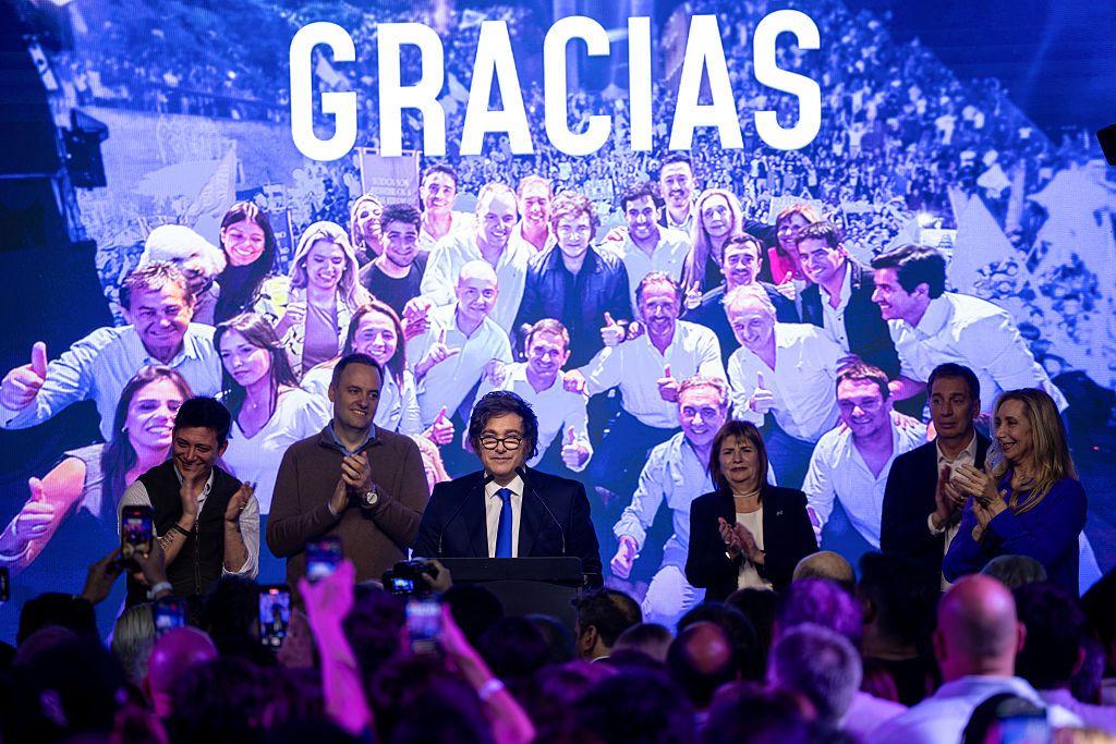 Milei e apoiadores posam para foto com painel ao fundo escrito Gracias