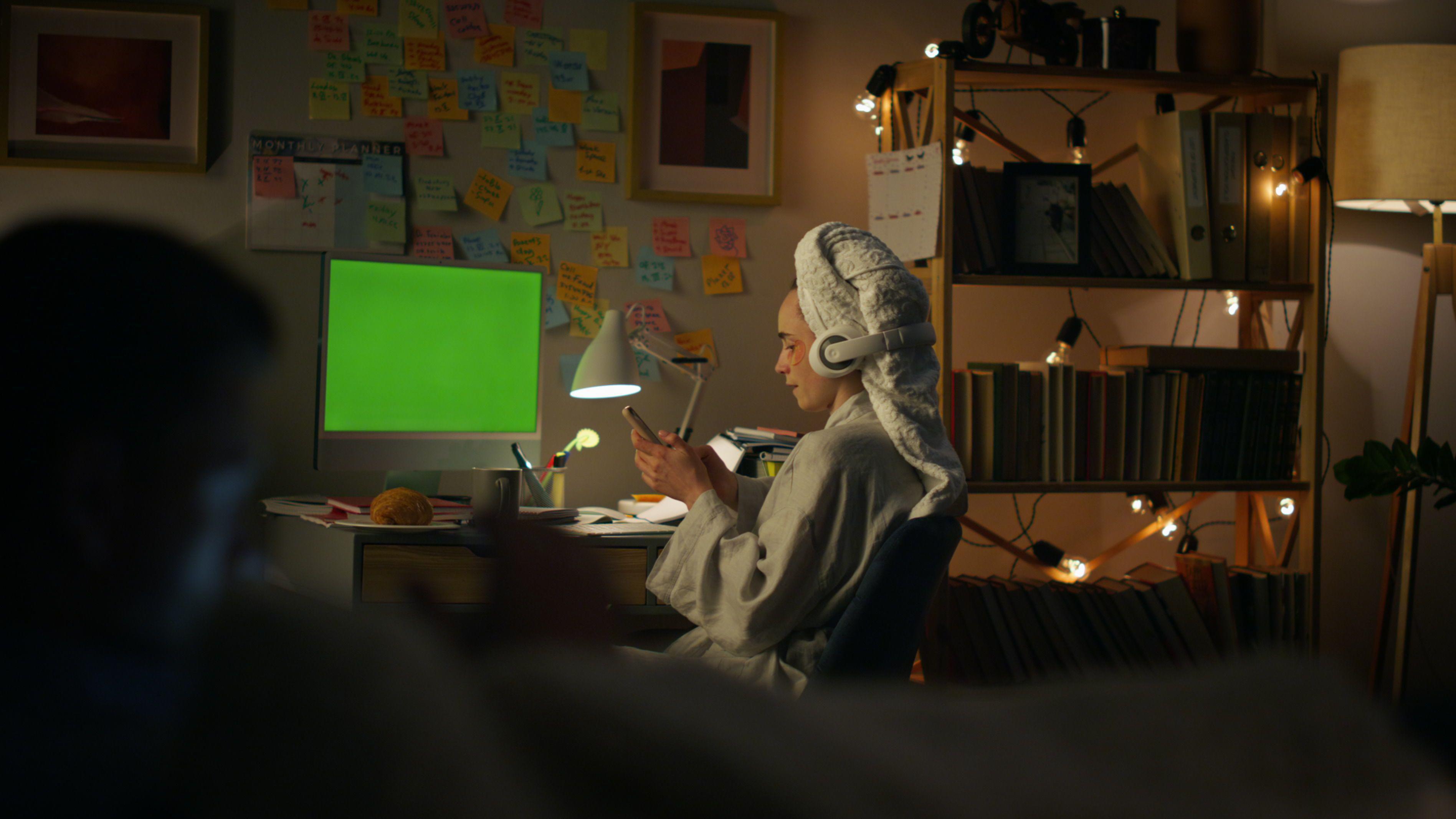 Mujer que se bañó por la noche, en una habitación iluminada solo por unas luces pequeñas, mirando el celular con una toalla en la cabeza y una computadora de fondo