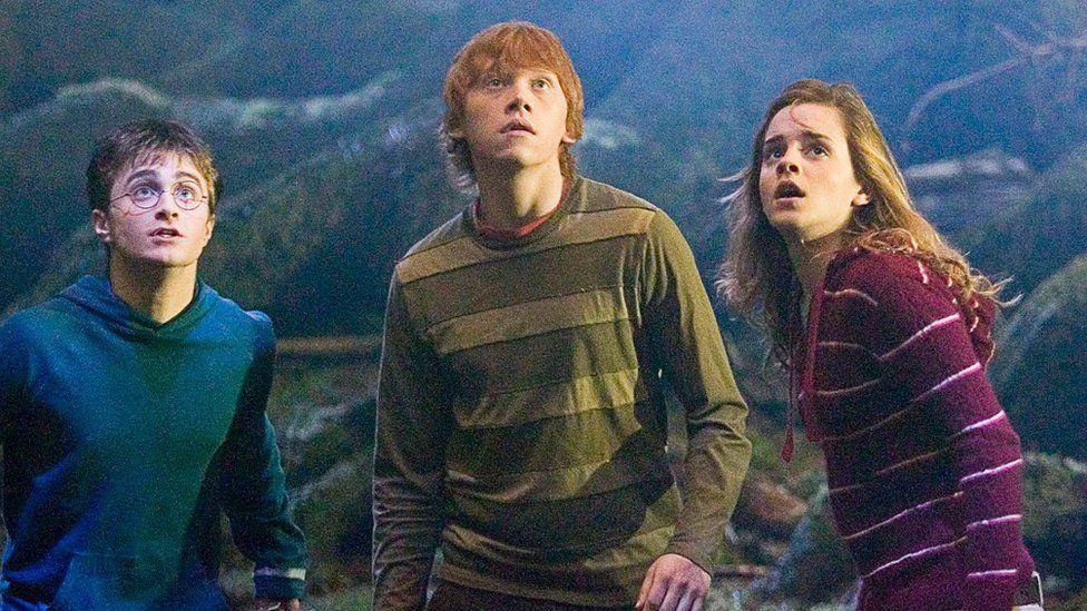 Da esquerda para a direita, Daniel Radcliffe (Harry Potter), Rupert Grint (Rony Weasley) e Emma Watson (Hermione Granger) aparecem em uma floresta, &agrave; frente de uma &aacute;rvore, com express&otilde;es de surpresa e apreens&atilde;o, olhando para algo acima deles. Radcliffe usa &oacute;culos redondos e um moletom azul, Grint veste uma blusa listrada em tons de verde e marrom, e Watson aparece com um casaco vinho com listras claras.