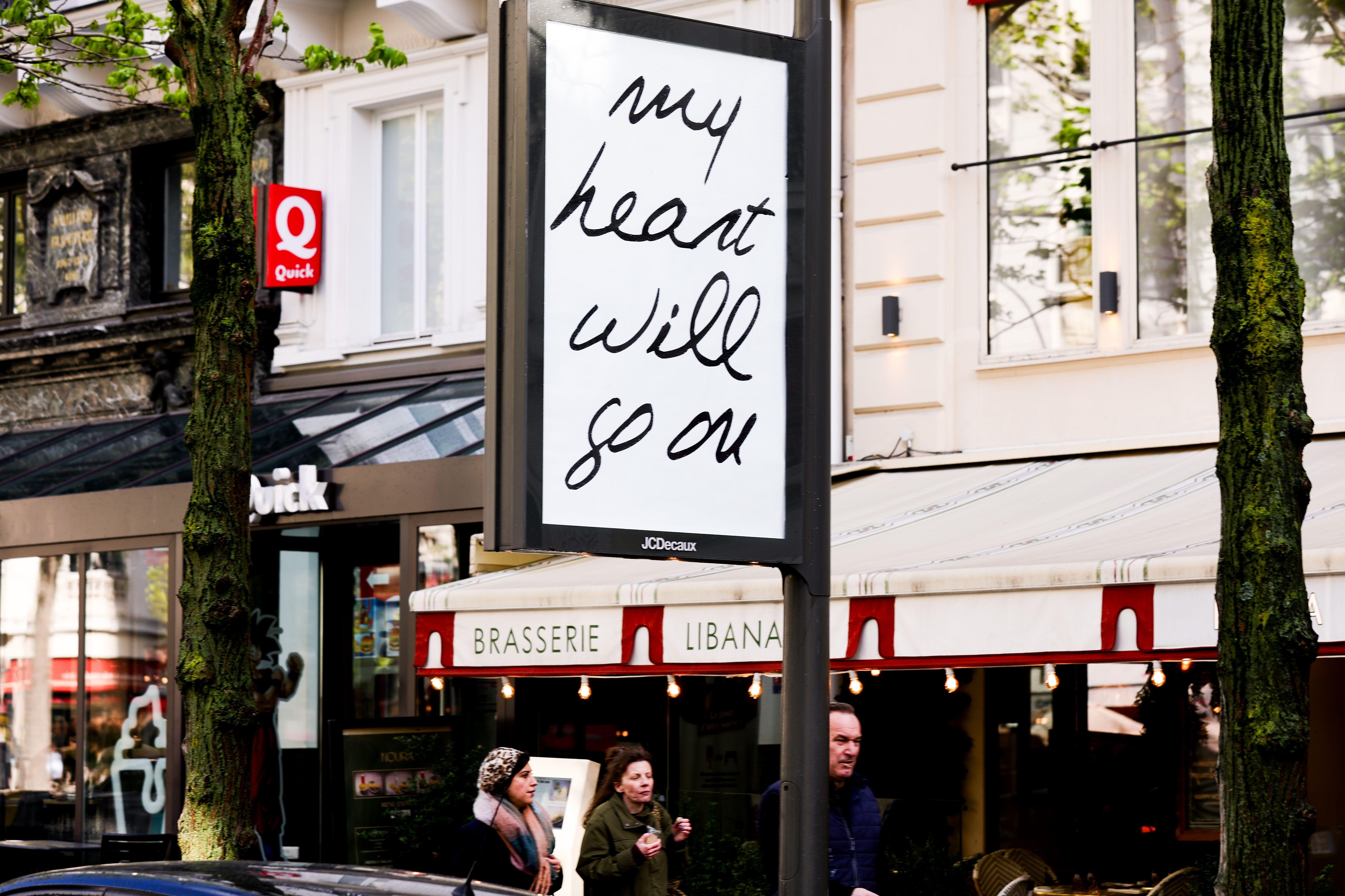 Um outdoor com a frase &ldquo;My Heart Will Go On&rdquo; aparece tendo ao fundo uma brasserie parisiense