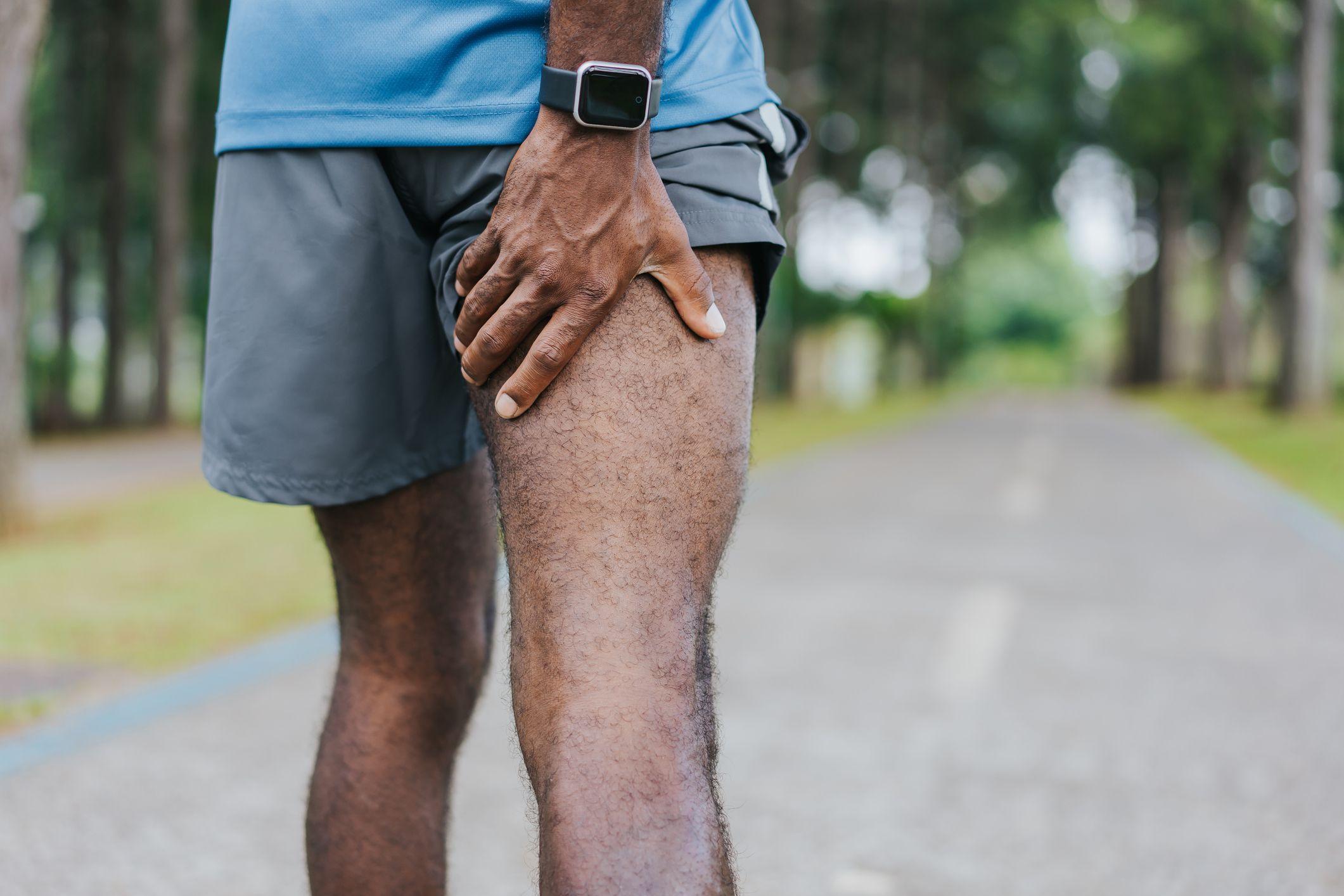 A close up shot of a man's thighs from behind. He is holding the back of his thigh with his hand. He is outside, and we can see blurry trees in the background. He is wearing gray shorts, a blue t-shirt and a smart watch. 