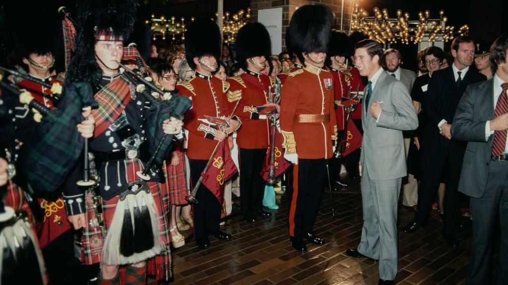 Princ u sivom odelu okružen grupom ljudi u crvenim uniformama Kraljeve garde