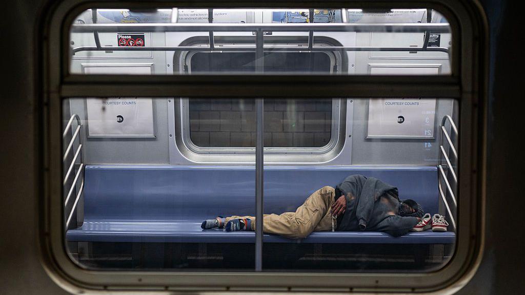 Homem sem-teto dormindo deitado no assento de um vagão parado na estação Jamaica Center do metrô de Nova York (EUA), uma das linhas mais usadas pelos sem-teto para refúgio na cidade, em Queens, Nova York, no dia 7 de abril de 2025