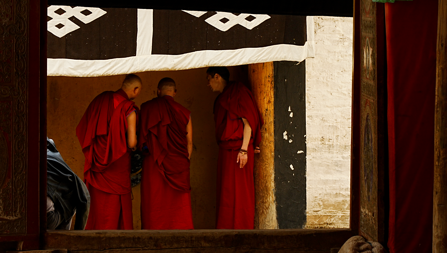 Tres monjes vestidos con túnicas rojas están de pie en fila, hablando entre ellos, con el rostro apartado de la cámara.