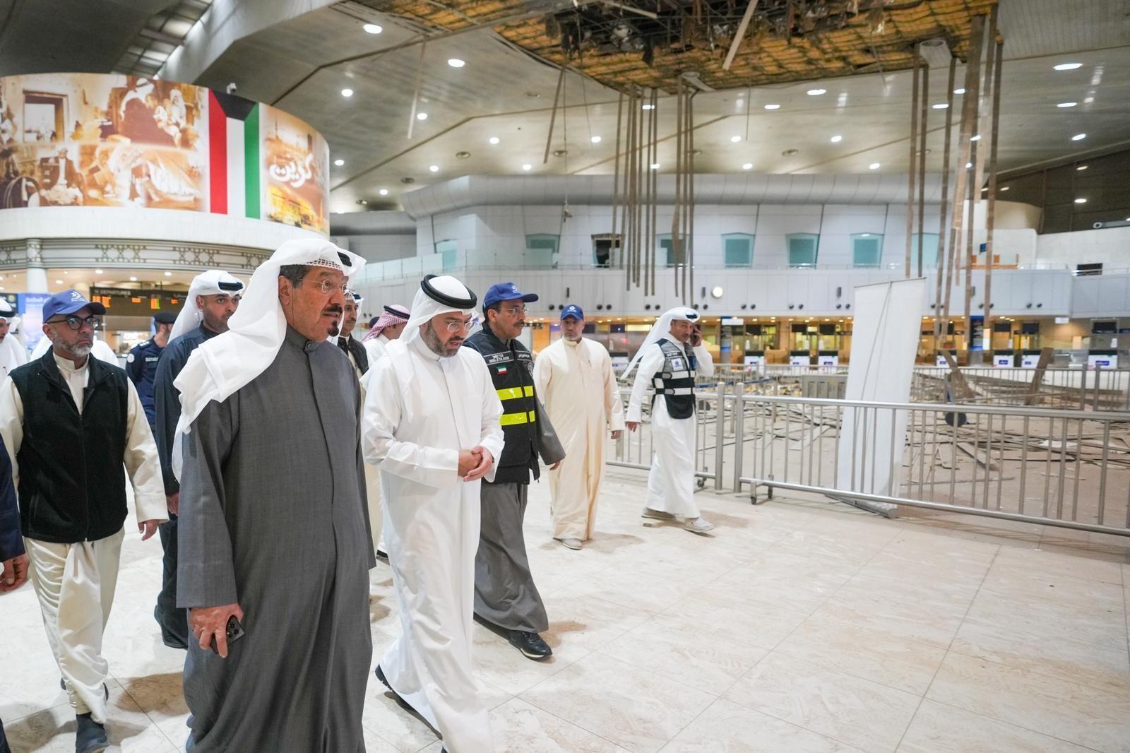 Ahmad al Abdullah al Sabah camina con otros dignatarios por el edificio terminal del aeropuerto, junto a una zona acordonada donde se observan daños. El techo presenta deterioros, con baldosas retiradas y estructuras colapsadas.