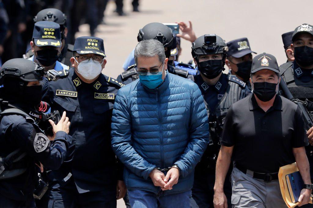 El expresidente de Honduras Juan Orlando Hernández, con un abrigo azul de plumas, lentes oscuros y esposado, es escoltado por miembros de las Fuerzas Especiales de la Policía para ser extraditado a Estados Unidos para enfrentar cargos relacionados con el narcotráfico, en la Dirección Nacional de Fuerzas Especiales de Honduras el 21 de abril de 2022 en Tegucigalpa, Honduras. (Foto de Jorge Cabrera/Getty Images)