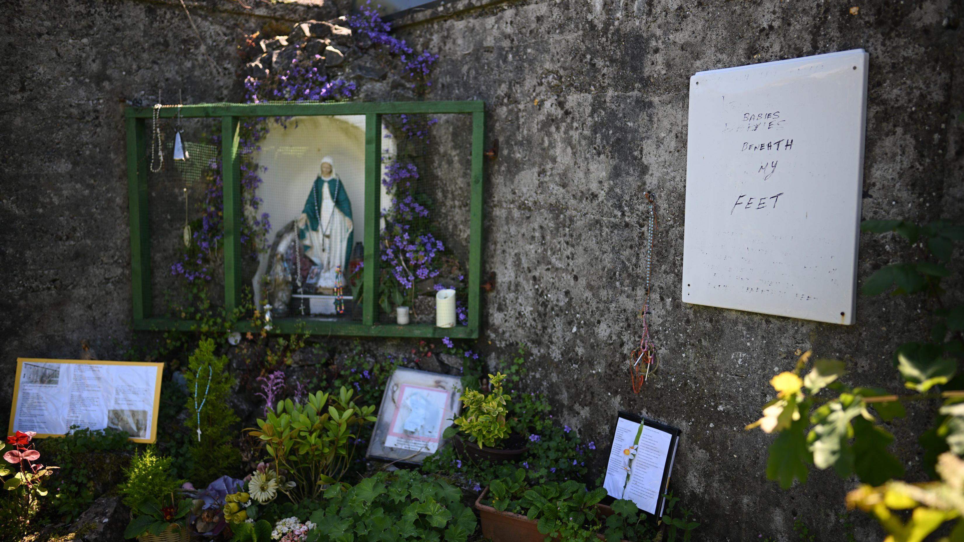 Dos altos muros de piedra se cruzan en ángulo recto en un jardín. En la esquina del ángulo se encuentra una gruta centrada en una estatua de la Virgen María. Está rodeada de flores y arbustos. Hay velas en un estante cerca de los pies de la estatua. También hay mensajes debajo de la estatua dejados por la gente y, a la derecha, una pizarra blanca con un mensaje de recuerdo.