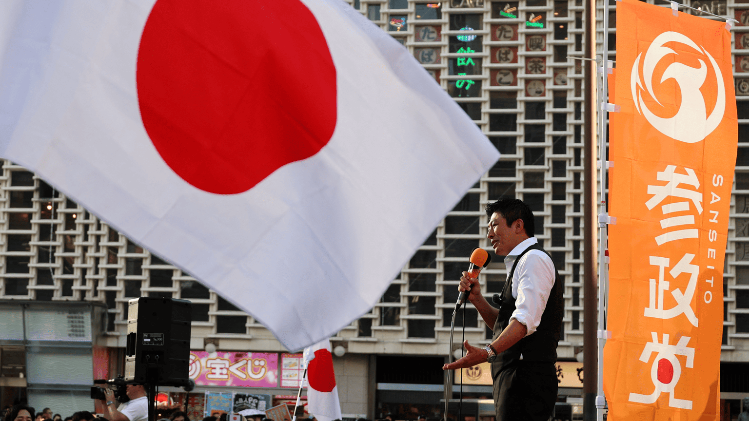 日本參政黨黨魁神谷宗幣在該黨集會上發表演說，畫面前景是一面大型旗幟，神谷身穿白襯衫，手持麥克風。