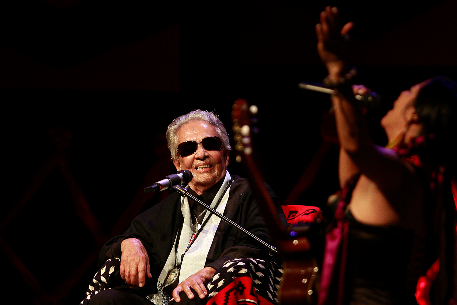 Chavela Vargas, con lentes oscuros y una sonrisa, está sentada con un micrófono al frente. Ve a Lila Downs quien canta con un micrófono y sube uno de sus brazos