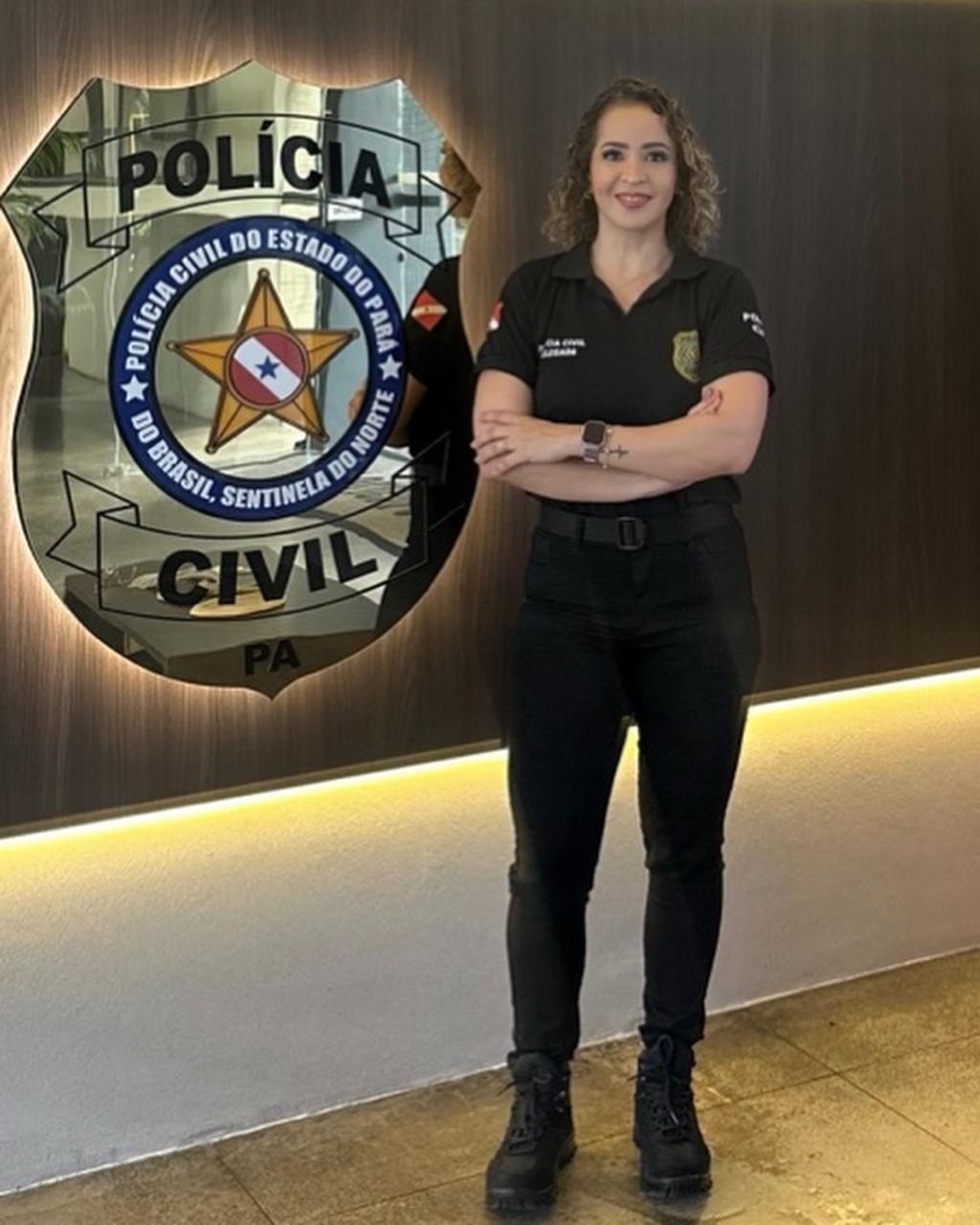 A delegada Amanda Souza o lado de um logotipo da Pol&iacute;cia Civil. Ela veste roupas pretas da pol&iacute;cia e cal&ccedil;a bota e est&aacute; com os bra&ccedil;os cruzados, sorrindo de p&eacute; para a c&acirc;mera