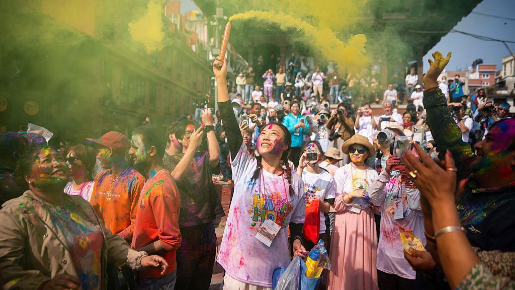 Žena namazana prahom po licu ispaljuje dimnu bombu tokom proslave festivala, a oko je nekoliko ljudi 