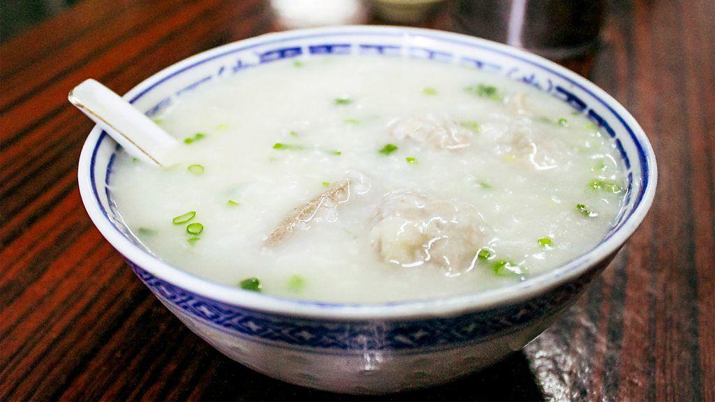 Bol de congee con cebollino y trozos de carne o dumplings, servido con cuchara de cerámica sobre mesa de madera.