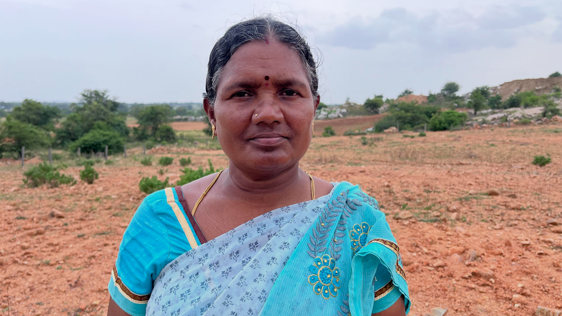 Seorang perempuan berusia 50 tahun mengenakan sari berwarna biru kehijauan bermotif bunga dan daun. Rambut gelapnya diikat ke belakang dan ia mengenakan kalung serta anting emas. Di belakangnya terdapat tanah kosong dan beberapa semak belukar.