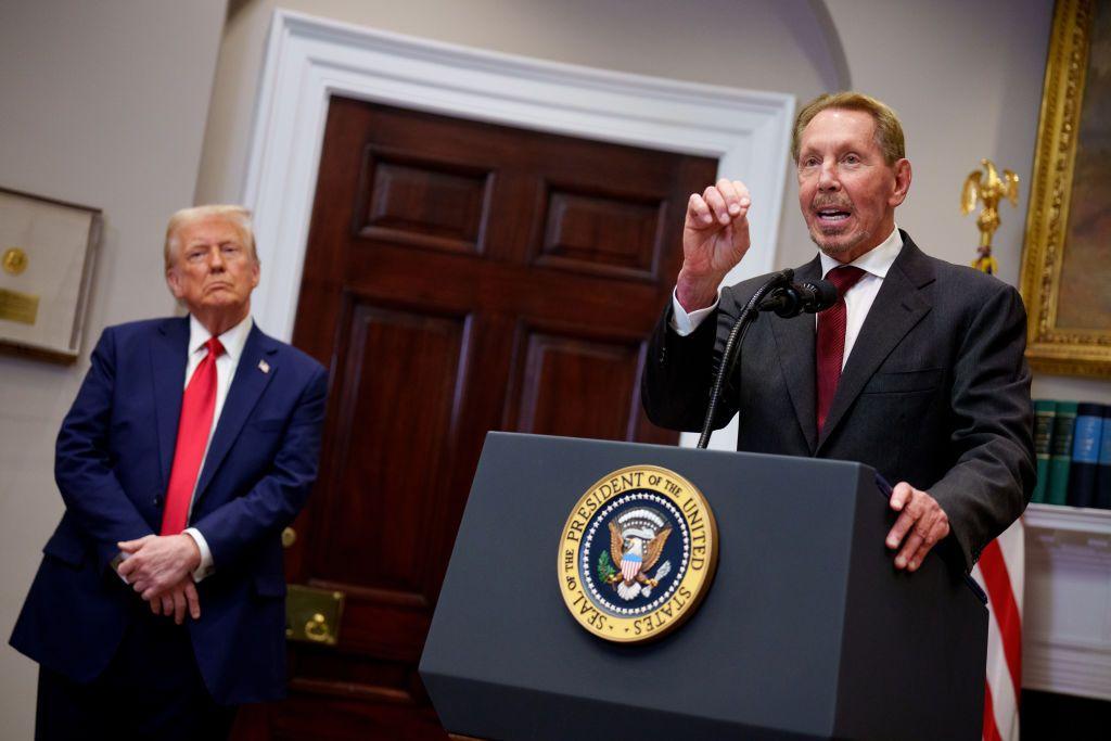 Larry Ellison, a la izquierda, vestido con traje oscuro, camisa blanca y corbata roja, habla desde un podio en el Despacho Oval. A su izquierda, Donald Trump con traje azul, camisa blanca y corbata roja, le escucha y observa.