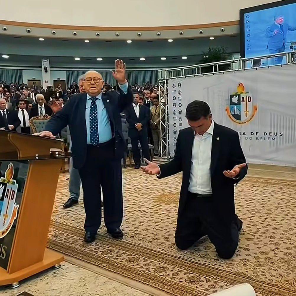 Fl&aacute;vio Bolsonaro, de roupa social, ajoelhado e olhando para o ch&atilde;o, durante culto evang&eacute;lico, ao lado de um pastor que discursa para os fi&eacute;is com um bra&ccedil;o erguido