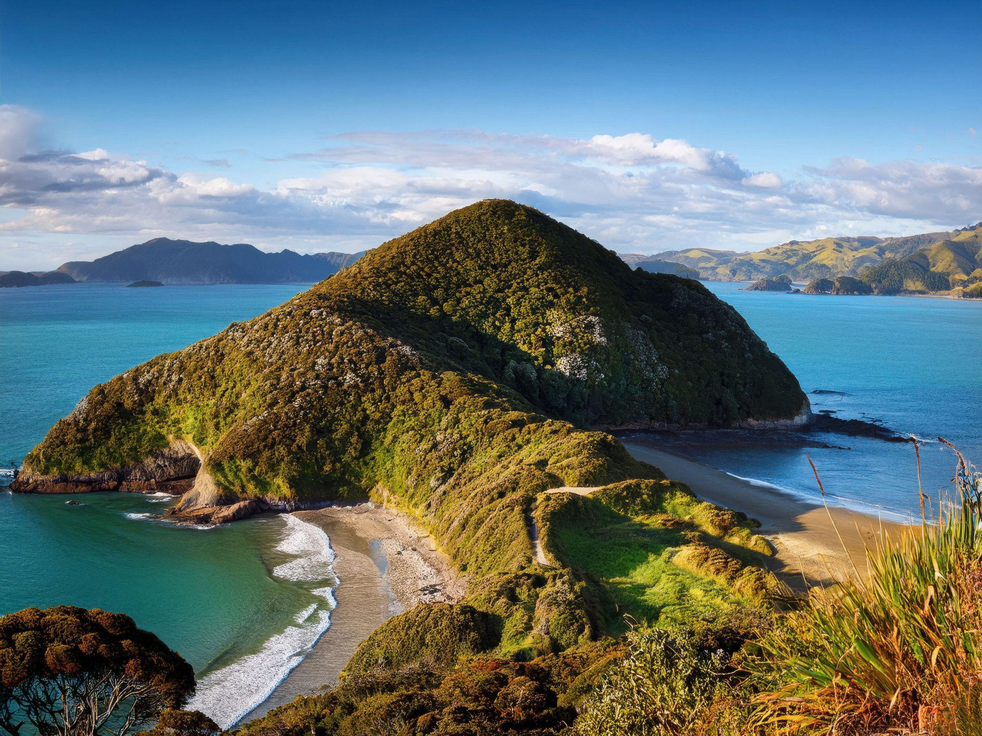 Vista aérea de uma costa montanhosa exuberante e verdejante com águas oceânicas turquesa sob um céu azul, na Ilha dos Macacos, Nova Zelândia