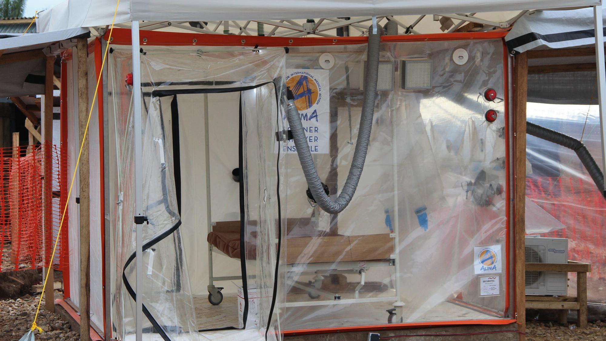 Uma estrutura transparente de pl&aacute;stico em formato de cubo, com uma porta de z&iacute;per aberta. H&aacute; uma cama vazia no interior e nenhum paciente