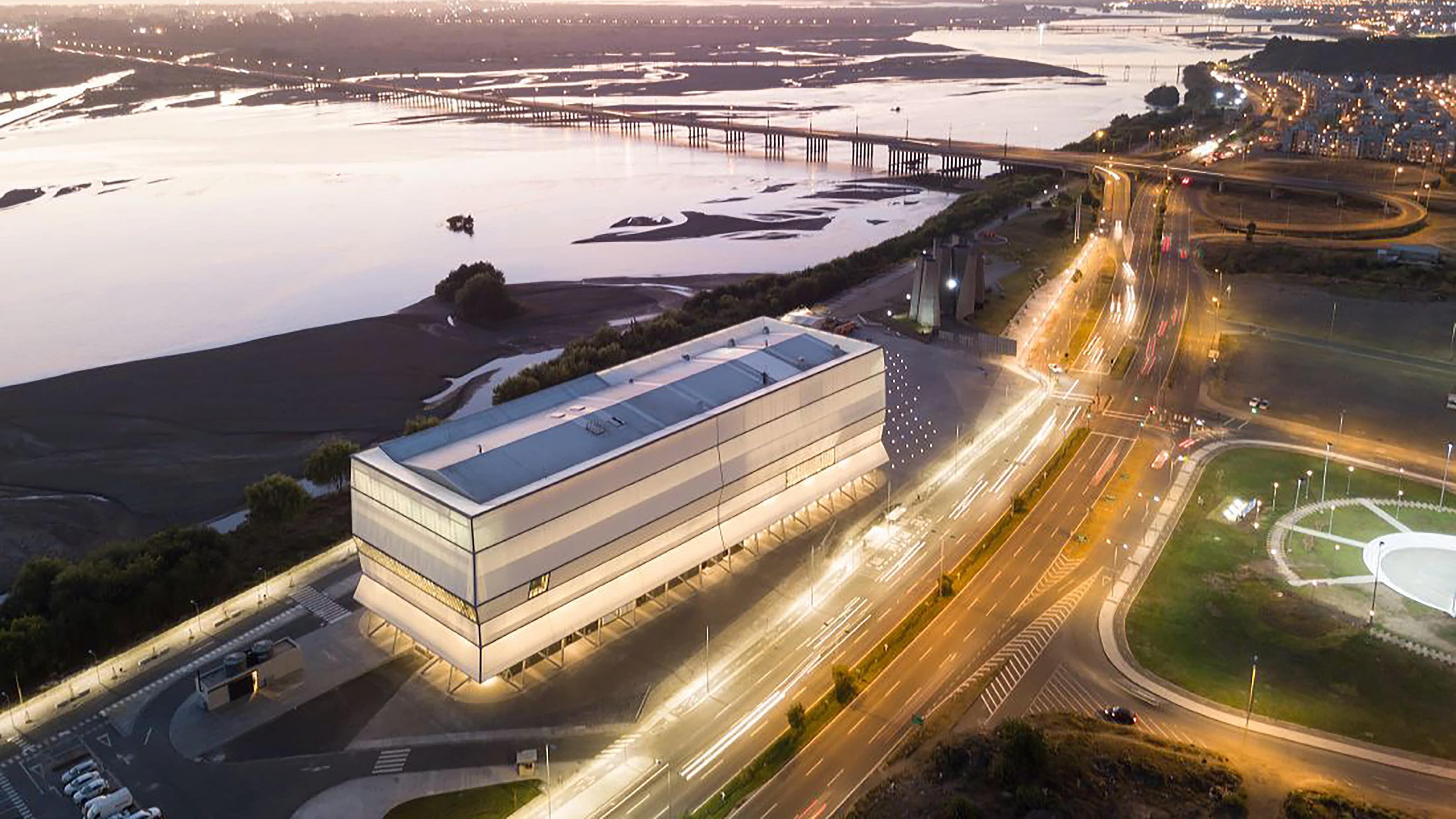 Edificio moderno junto a una carretera iluminada al atardecer, con un río amplio y puentes al fondo.