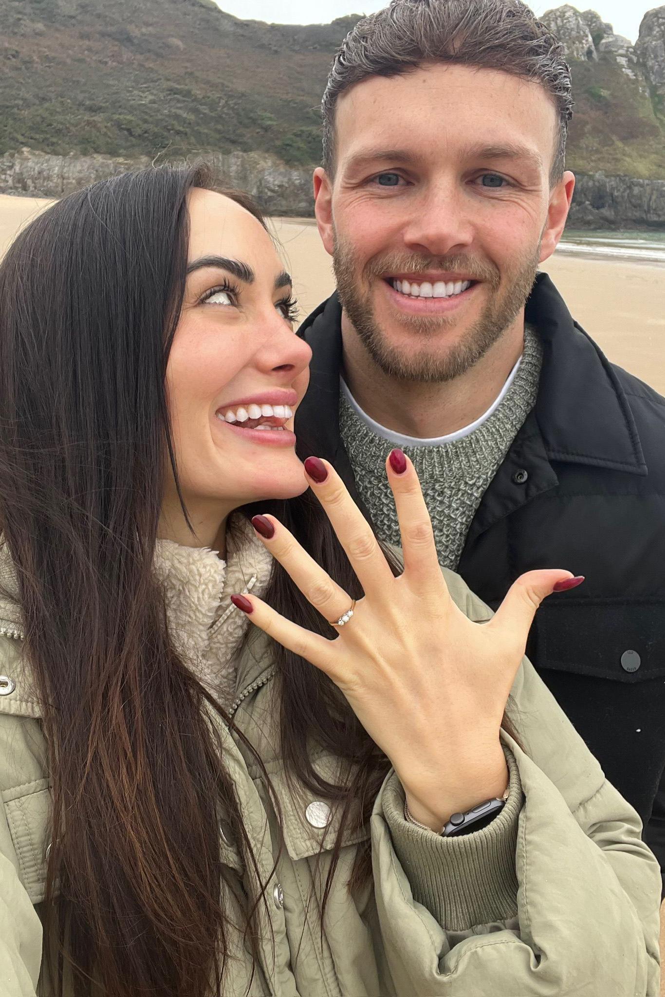 Lauren Macpherson e seu noivo, Zak. Ela levanta a m&atilde;o, com as unhas pintadas de vermelho e um anel de noivado com tr&ecirc;s diamantes no dedo anelar. Lauren olha para Zak e sorri. Zak tem cabelo castanho e olhos azuis e sorri para a c&acirc;mera