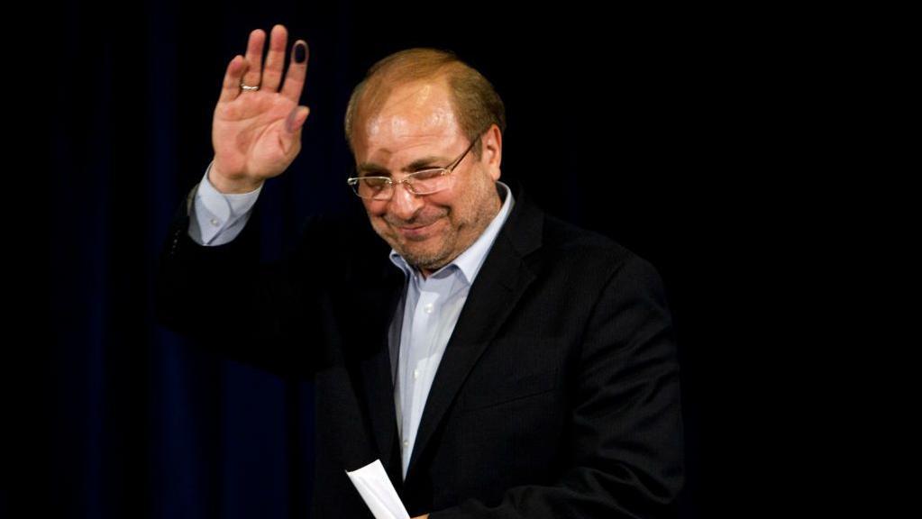 Mohammad Bagher Ghalibaf wearing a white shirt and black suit, waving and standing against a black background.