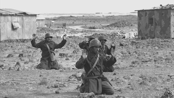Syrian soldiers kneel and raise their hands in surrender in the Golan Heights, on 10 October 1973, days into the Yom Kippur War. They are in uniform and wearing helmets. The ground is barren, with some shrubs growing. Behind them are several huts and a road winding into the distance.