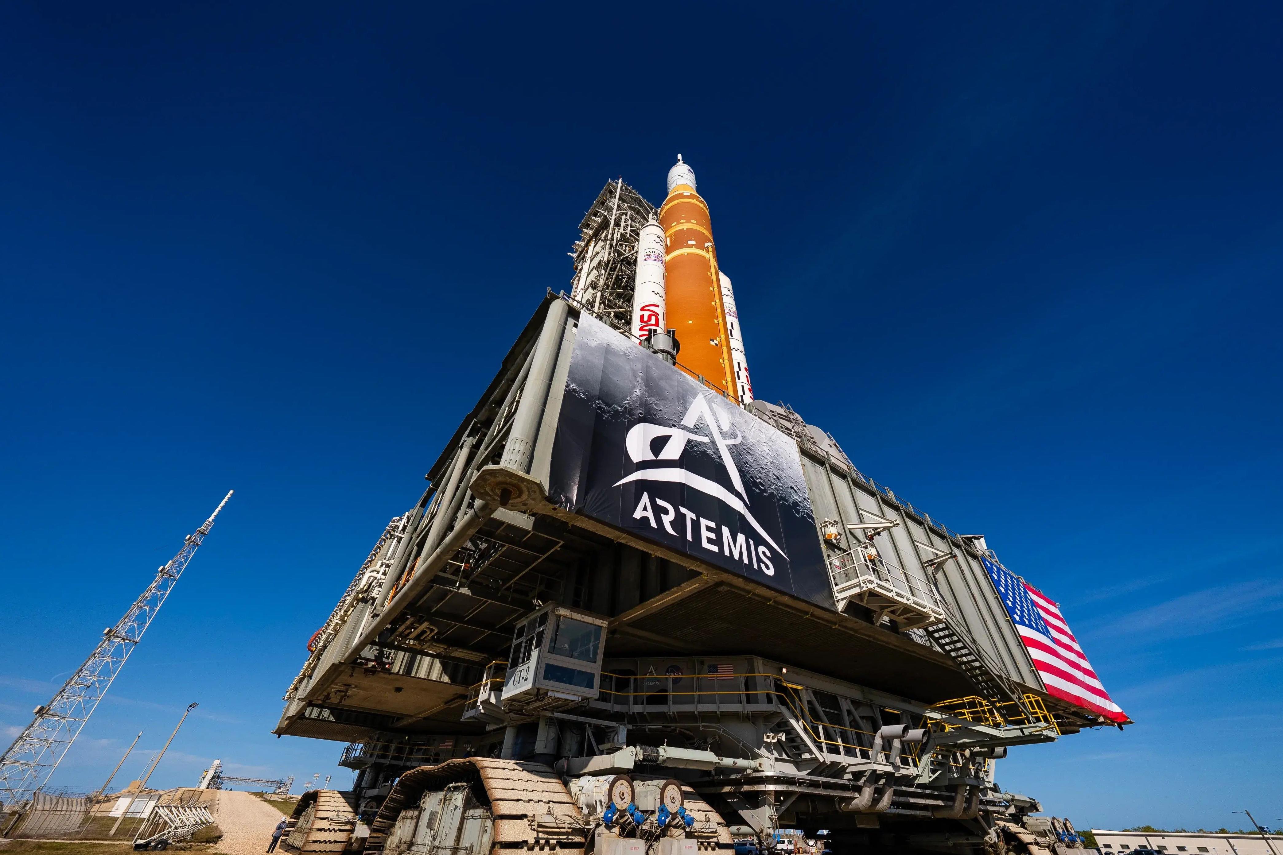 在佛羅里達州肯尼迪太空中心，NASA巨大的太空發射系統和獵戶座太空船在湛藍的天空下顯得格外醒目，底部懸掛著阿爾忒彌斯計畫的橫幅和美國國旗。