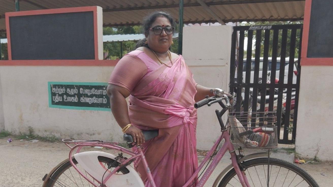 Jayachithra, vestindo um s&aacute;ri rosa e segurando uma bicicleta feminina rosa, em frente a uma casa