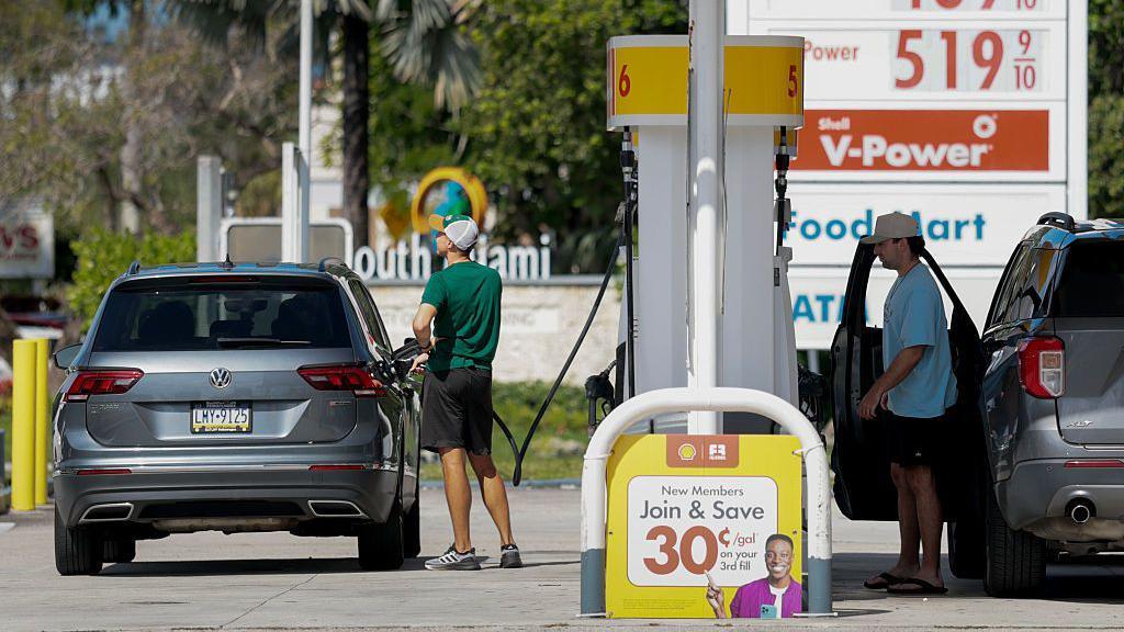 Dos conductores vestidos con pantalones cortos y camisetas llenan sus coches de gasolina.