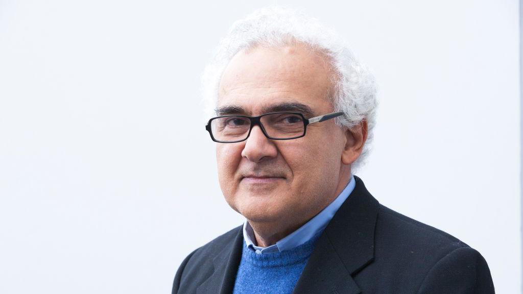 Milton Hatoum, um homem de cabelos brancos e &oacute;culos de arma&ccedil;&atilde;o preta, posa para um retrato. Vestido com blazer preto sobre um su&eacute;ter azul e camisa social clara, ele olha para a c&acirc;mera com express&atilde;o serena diante de um fundo branco.