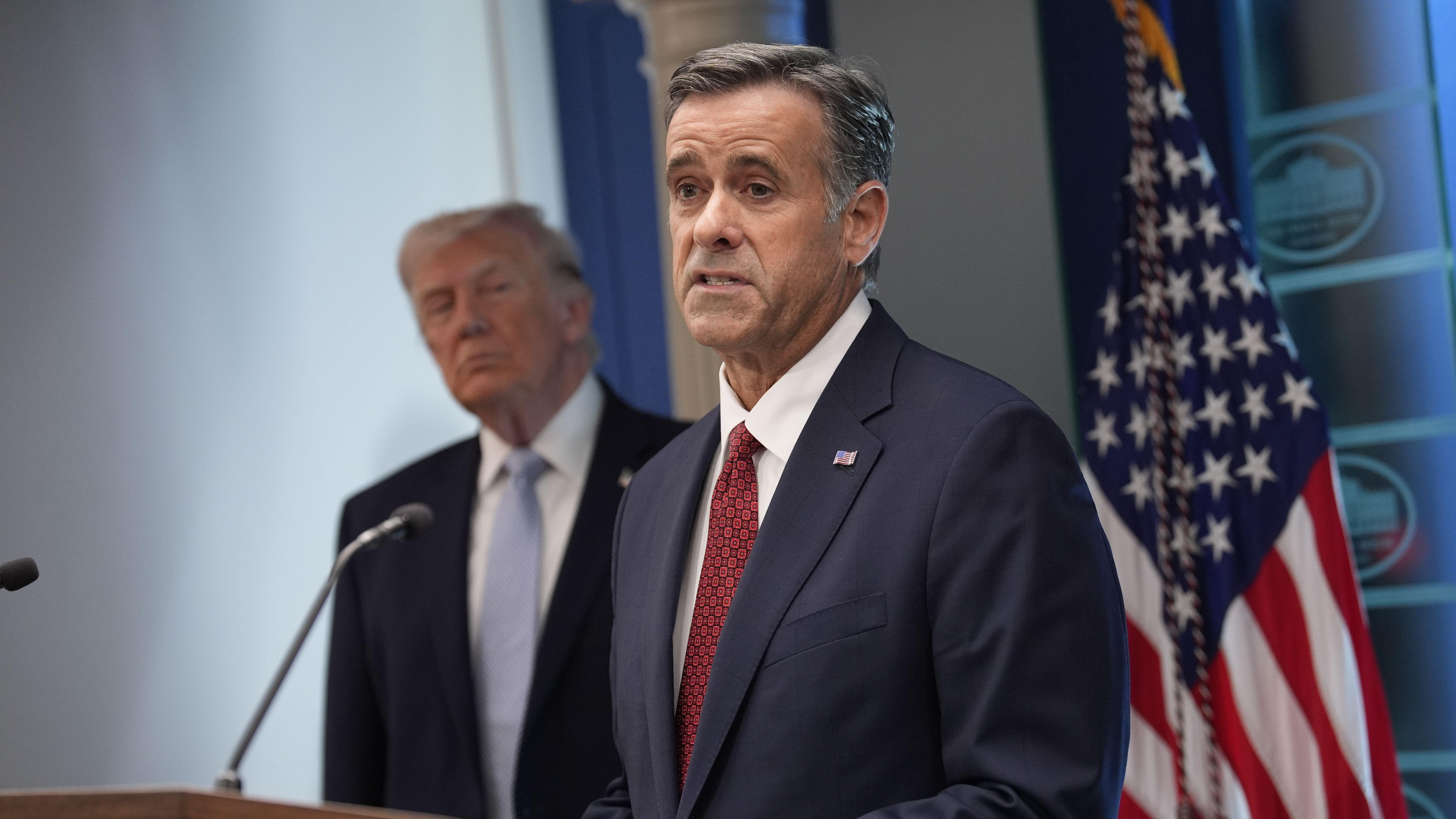 El director de la CIA, John Ratcliffe, hablando frente a un micrófono en la Casa Blanca, con Donald Trump detrás suyo. A su lado hay una bandera de Estados Unidos.