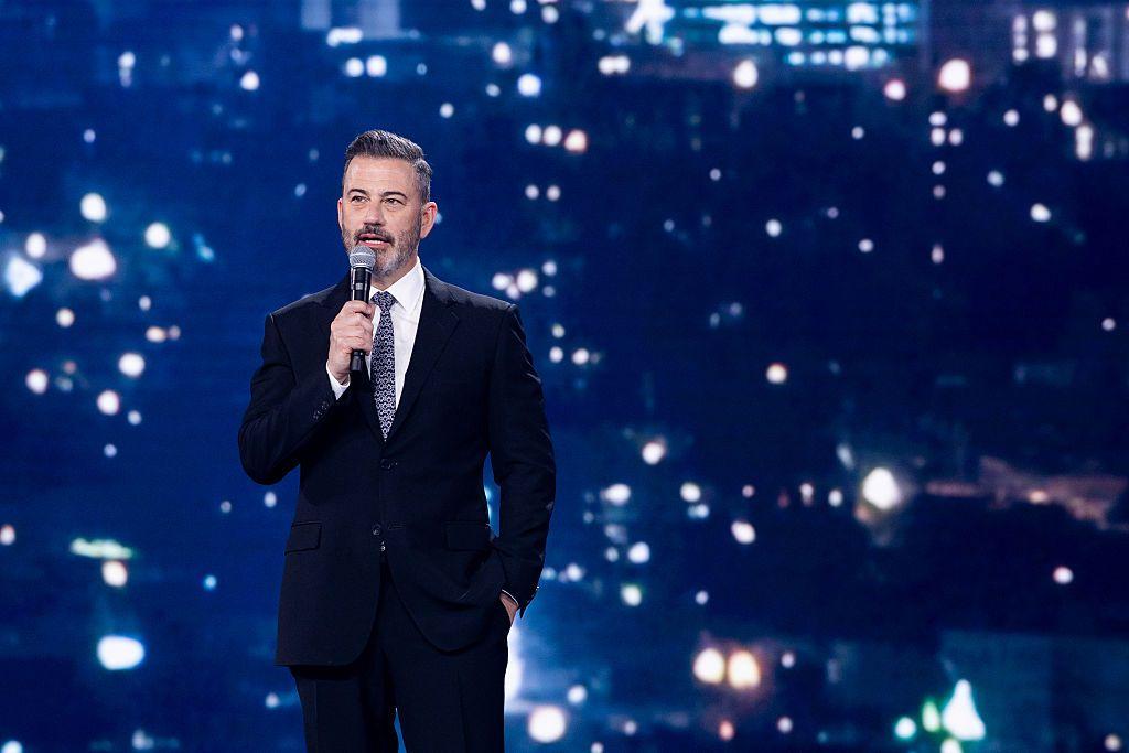 Jimmy Kimmel con un micrófono en mano y la otra mano en el bolsillo del pantalón. De fondo, luces borrosas y difuminadas
