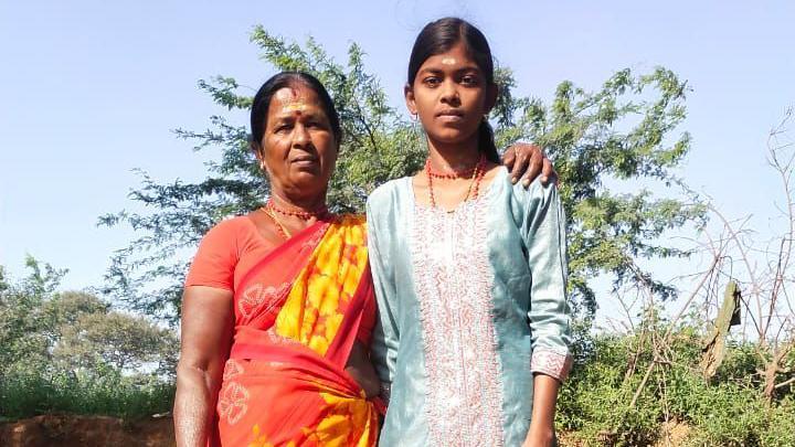 Vasantha veste um s&aacute;ri vermelho com estampa floral amarela, de p&eacute;, com a m&atilde;o no ombro da sua neta Naveena, que veste um top azul, com arbustos ao fundo