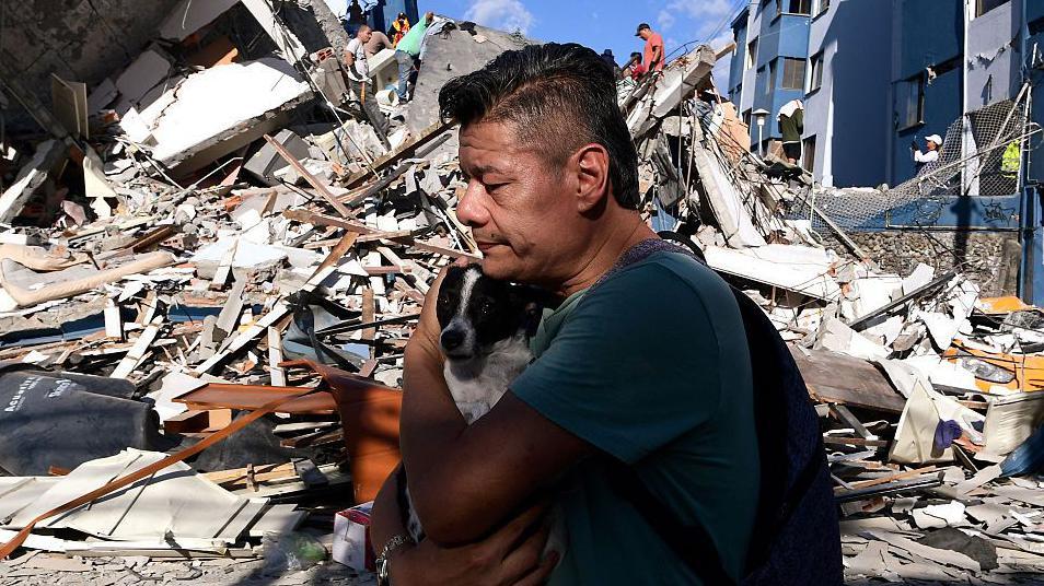 Un hombre abraza a una mujer y detrás los escombros de un edificio derruido.