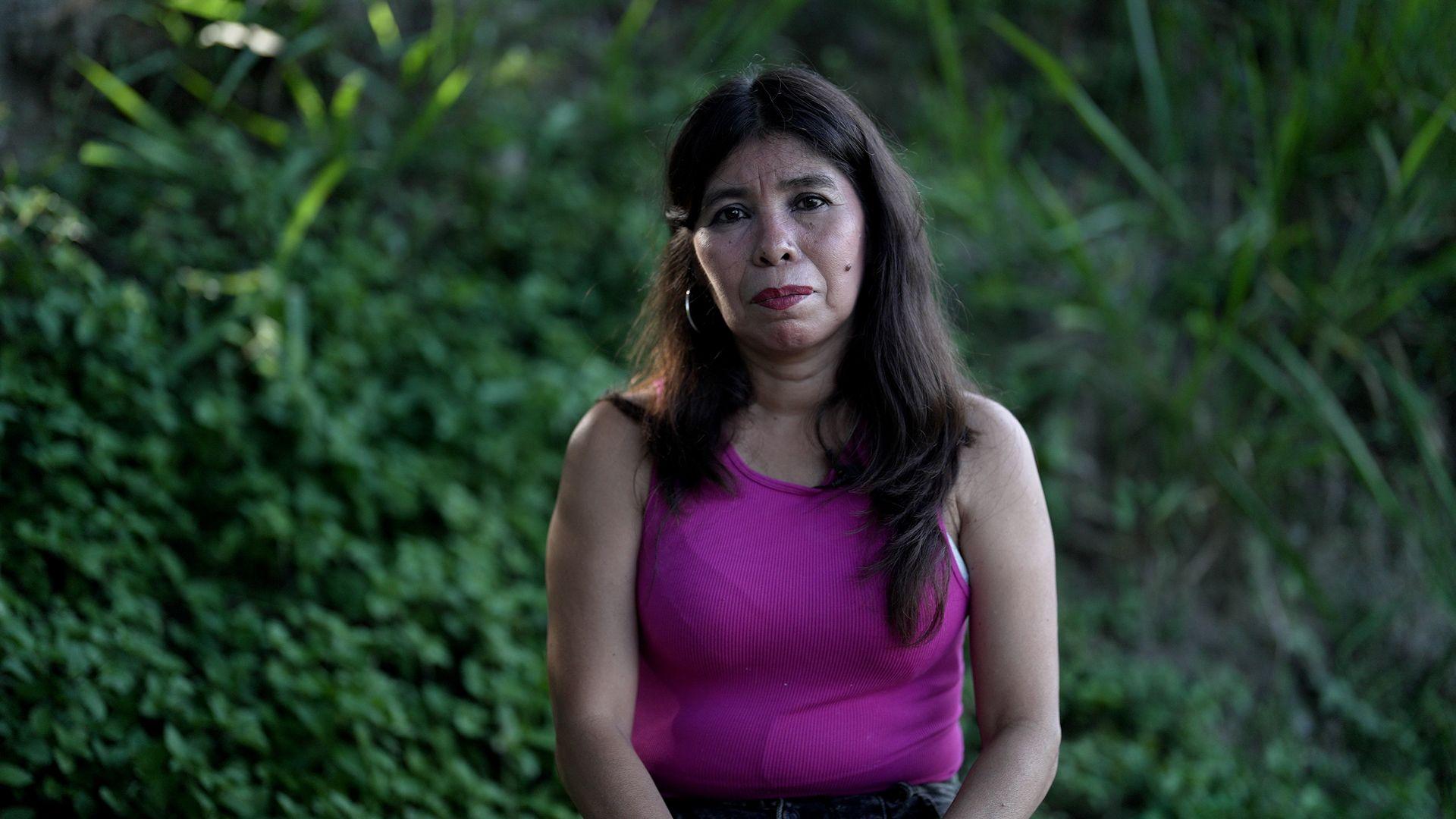 Mulher com blusa rosa com plantas ao fundo.