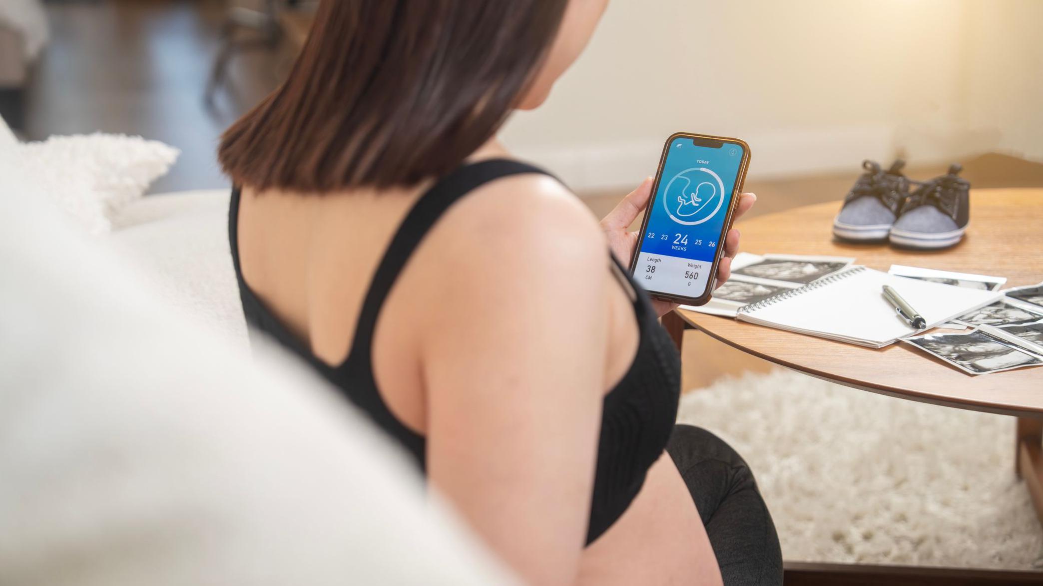 Una mujer embarazada consulta una aplicación en su teléfono móvil que contiene una ilustración sencilla de un feto con un cordón umbilical y unos números indicando las semanas que lleva embarazada