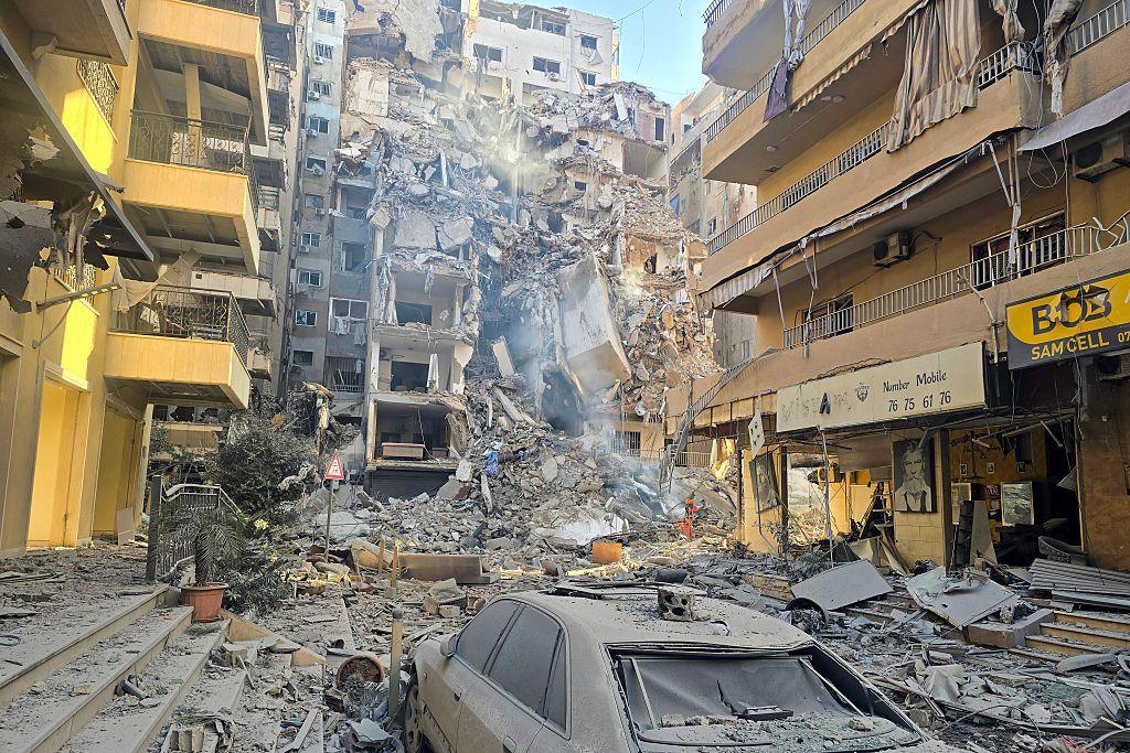 مبنى مدمر وسط مبانٍ أخرى، حيث تظهر آثار الدمار في الشارع، ويظهر في الصورة الملتقطة نهاراً تضرر إحدى السيارات.