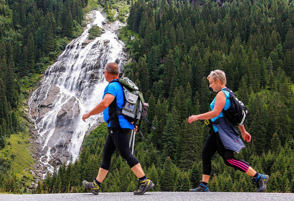 Una pareja de mochileros, con camisas azules y zapatos de montañistas, camina frente a una cascada que cae entre una empinada montaña con árboles verdes