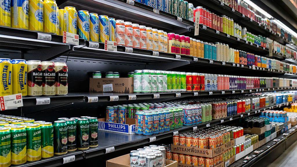 Prateleiras de supermercado repletas de latas de bebidas, com diferentes marcas, sabores, cores e embalagens, destacando a variedade dispon&iacute;vel no mercado.