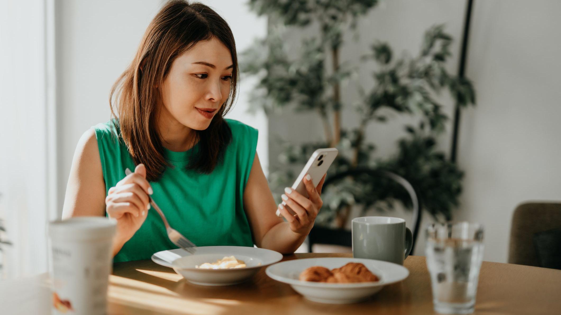 Mulher com traços asiáticos olhando o telefone celular enquanto come