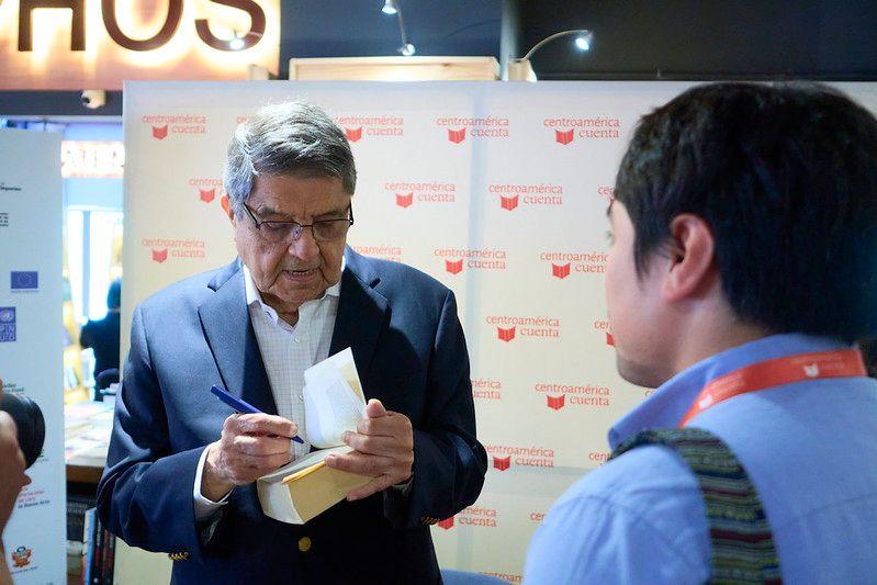 Una foto del escritor Sergio Ramírez autografiando un libro. Frente a él está un hombre que se ve de espaldas a la cámara. 