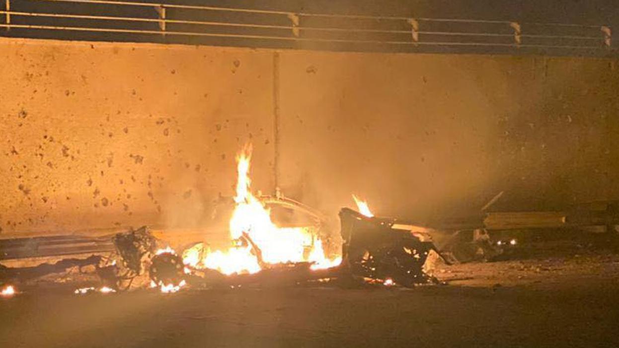 A handout photo made available by Iraq's military-run Security Media Cell via Facebook shows the remains of a burning vehicle beside a tall concrete wall. The hulk of wheels and the bonnet can be made out, with the main bulk of the vehicle in flames. The tall wall with railings on the top is behind the vehicle. It is night and orange flames illuminate the area.