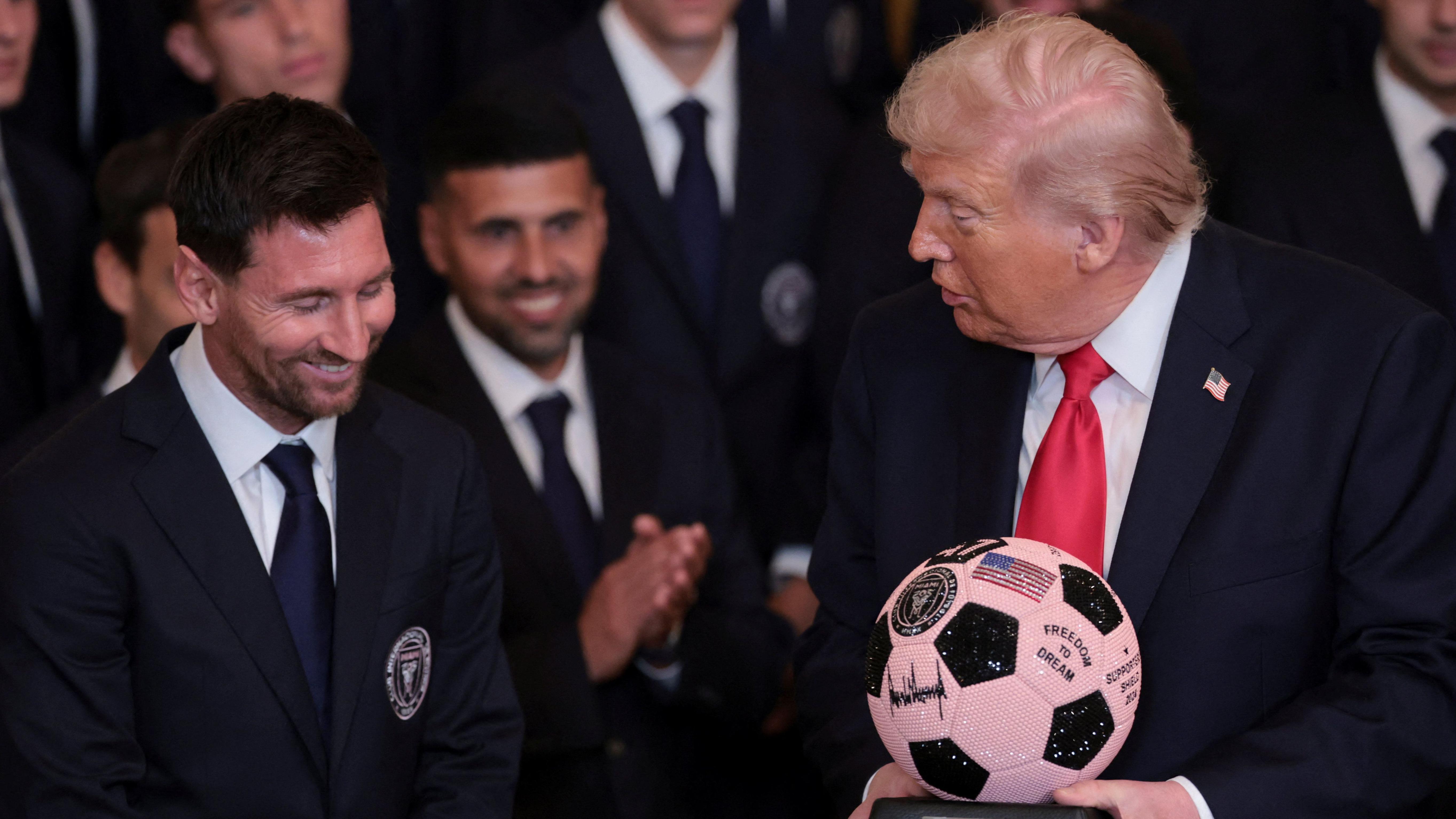 Lionel Messi, de camisa branca, terno azul escuro e gravata, sorri para o presidente americano Donald Trump, com roupas similares e gravata vermelha e a bandeira dos Estados Unidos na lapela, segurando uma bola de futebol oferecida de presente a ele durante uma recep&ccedil;&atilde;o para os jogadores do Inter Miami na Casa Branca, no dia 5 de mar&ccedil;o de 2026