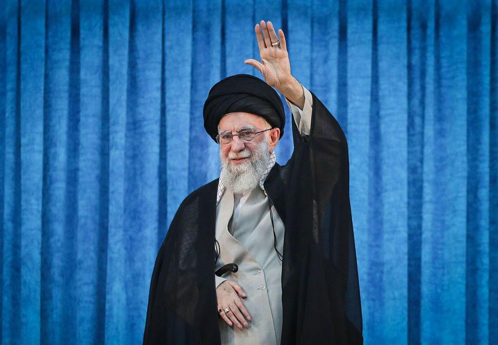 يقف المرشد الأعلى الإيراني السابق آية الله علي خامنئي، رافعاً يده اليسرى، أمام ستارة زرقاء في حفل أقيم في 4 يونيو 2025.
