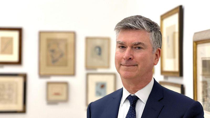 O advogado Christopher Marinello, de pé, em frente a duas paredes com diversos quadros