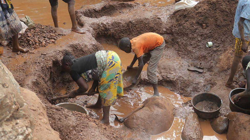 Two women - one in a yellow wrap skirt and dark top, the other in an orange T-shit and three-quarter length trousers - stand in muddy water with bowls as the search for gold.