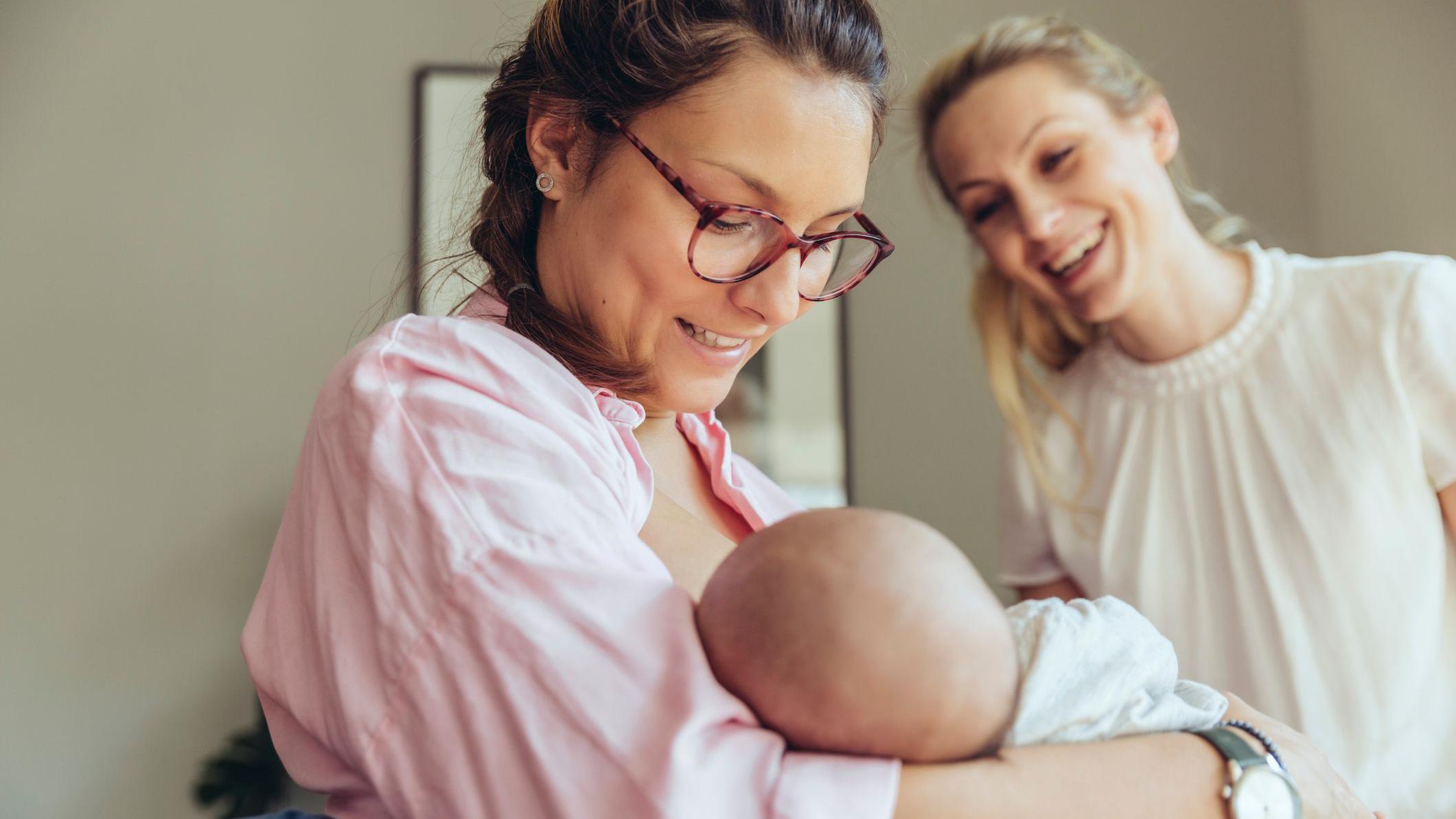 Una mujer con su bebé en brazos y otra que la mira sonriente.